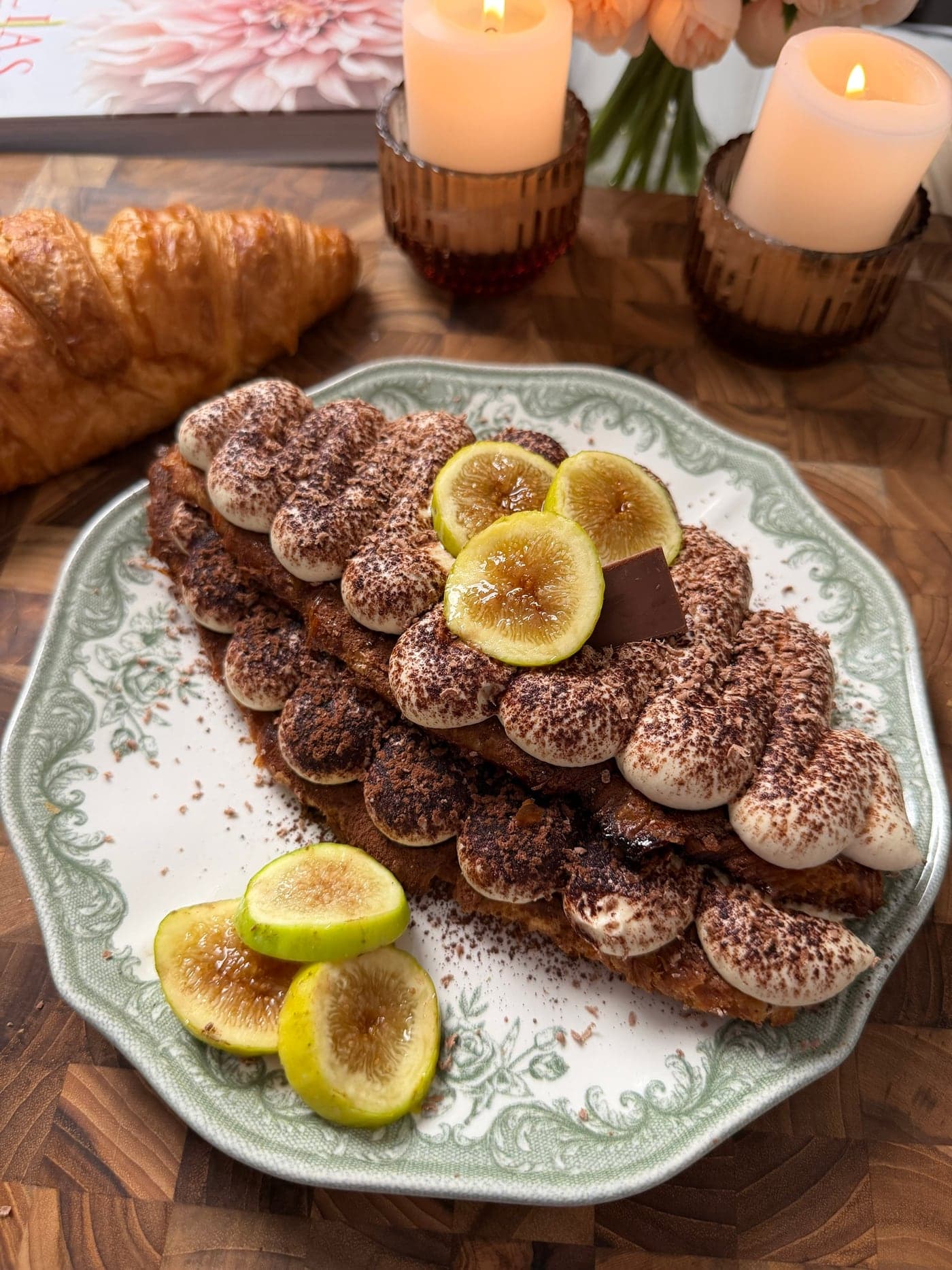 Croissant tiramisu layered with cocoa-dusted cream, sliced green figs and a square of dark chocolate on a green floral plate